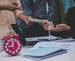 Hands exchanging money at a business desk with a laptop and clock.